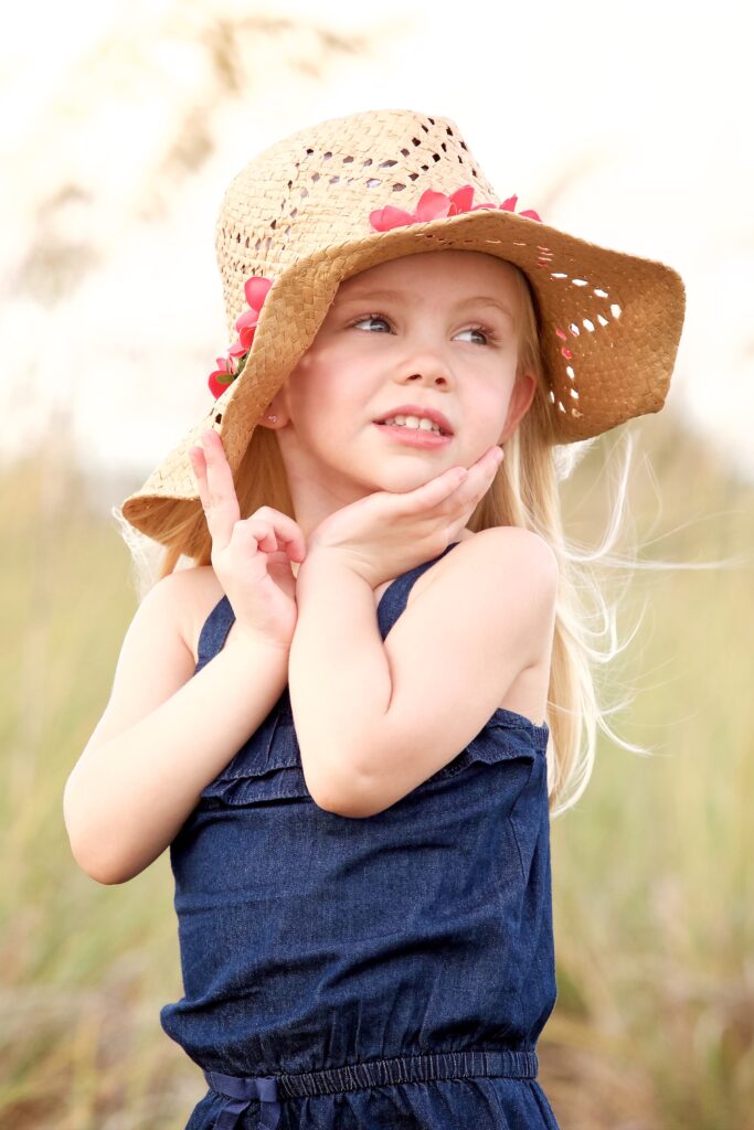 close up of toddler in a blue dress holding her sunflower hat over her ears