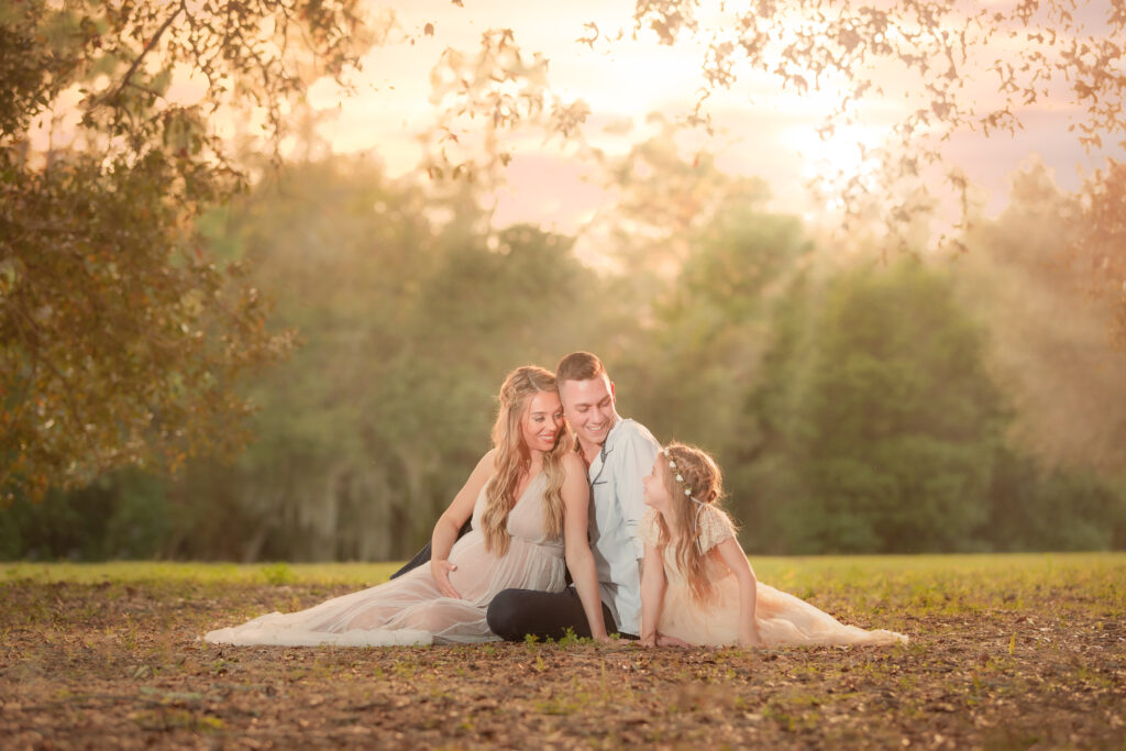 family of 3 sitting in the park for maternity photoshoot. Photo taken during golden hour so the sun is shining through the trees