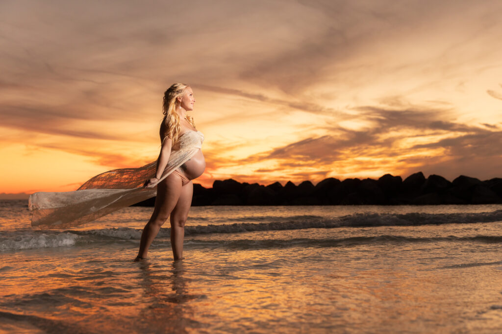 Pregnant woman poses in ivory lace dress at St. Pete Beach at sunset.