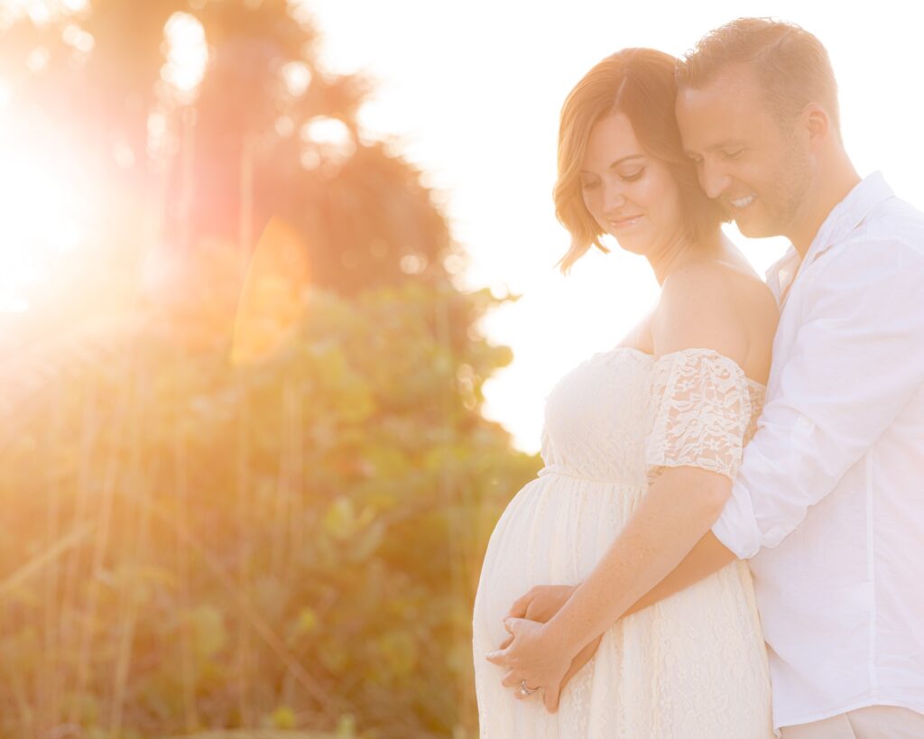 Pregnant woman in white dress is embraced by her husband