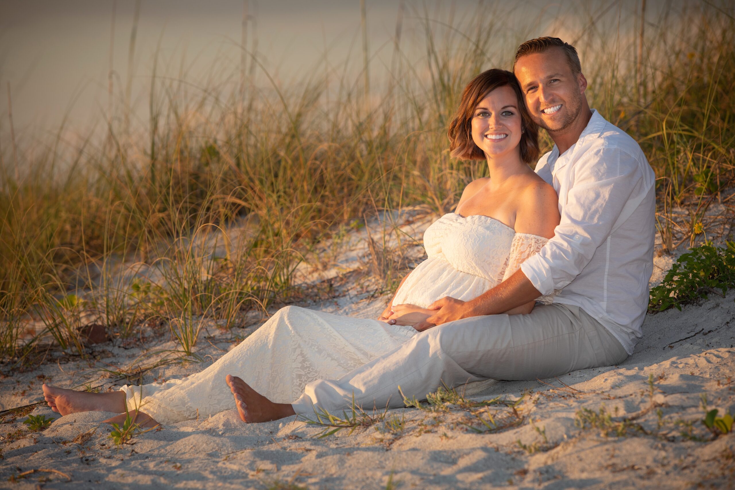 Pregnant couple sits in sand in white at Sand Key Beach
