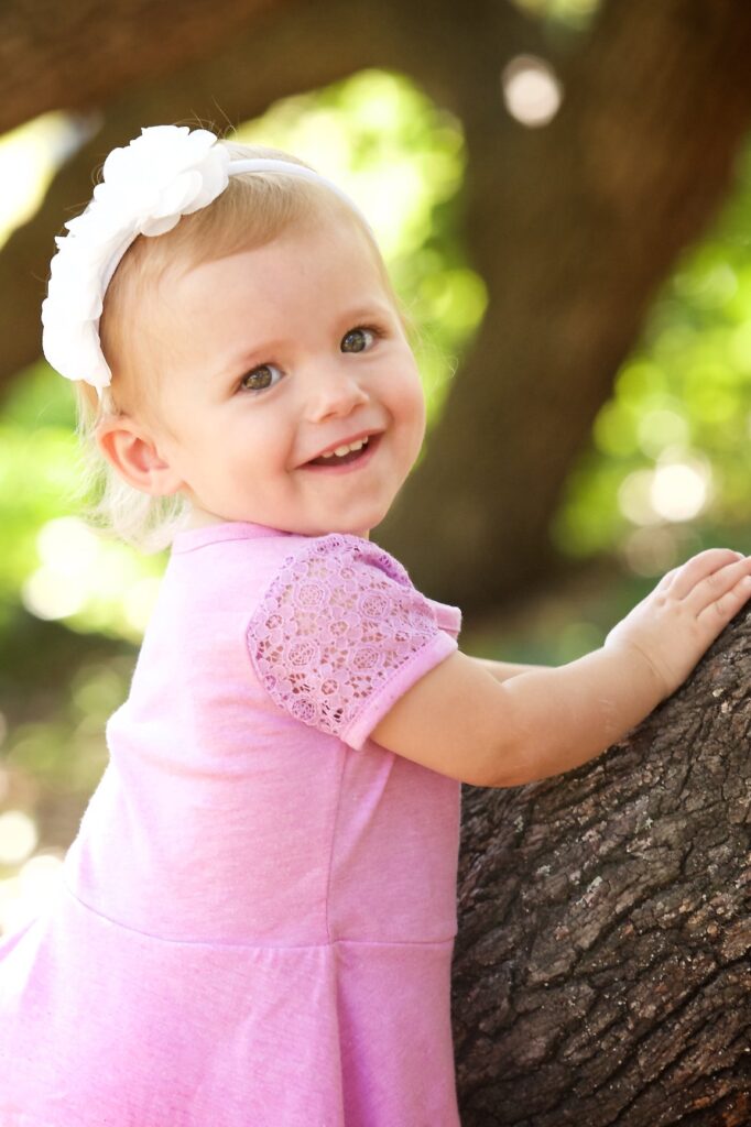 close up little girl  in a pink dress holding onto a tree branch smiling at the camera