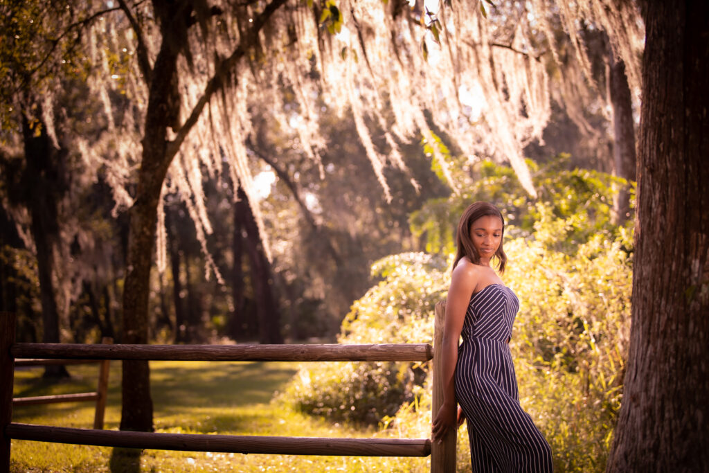 woman leaning against fence wearing a stripe jumpsuit. Light shining through oak tress in background at Lakes Park near Tampa