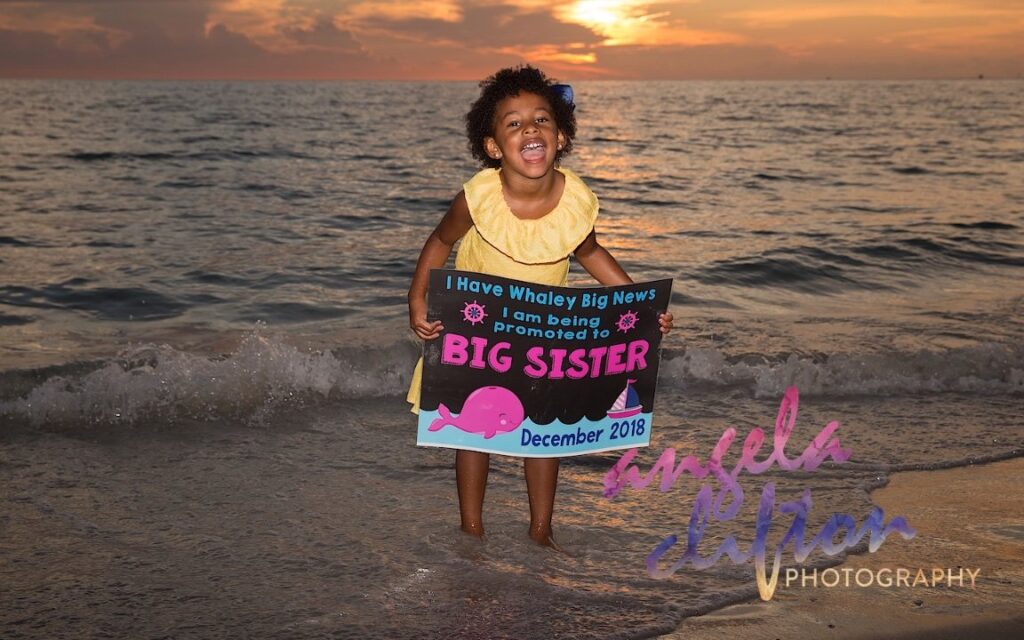young girl holding a sign that says "BIG SISTER." She's standing on a Florida beach with the sun setting in the background. She's excited!