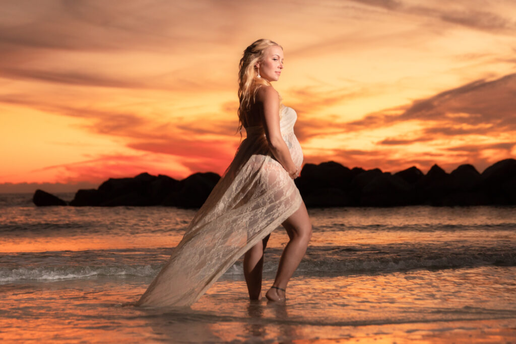 Sunset maternity photo shoot at park in clearwater - pregnant blonde woman wearing a white dress, standing on the shore against an orange sunset