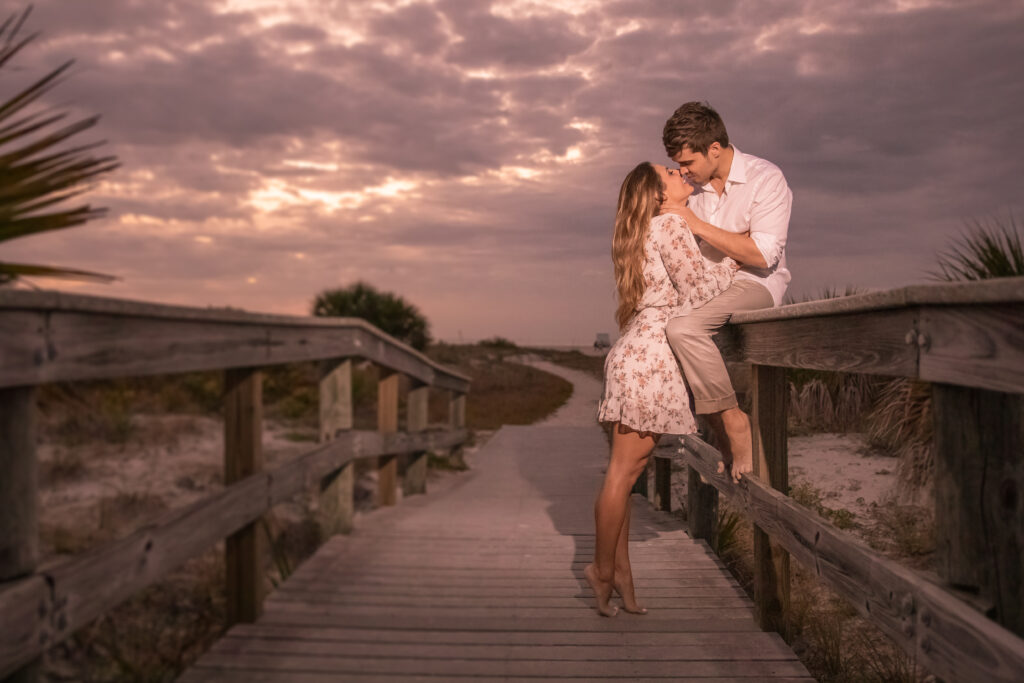 summer beach photoshoot idea: man sitting on a railing leaning down kissing his girlfriend/fiance/wife. There is a beach horizon in the background and the sun is peeping between the clouds