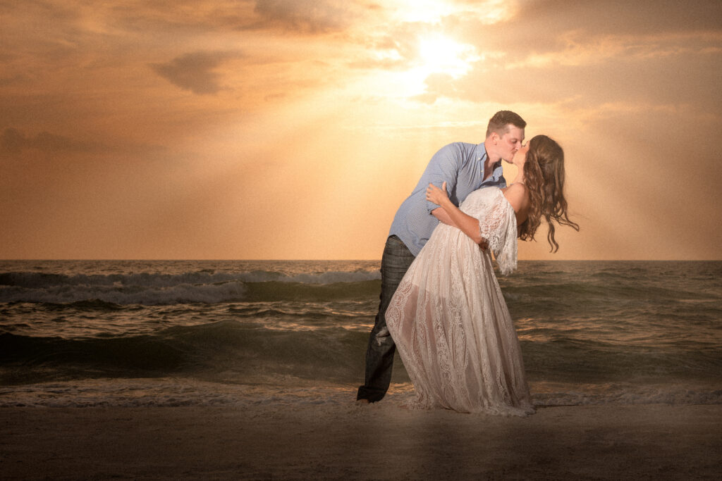 Angela Clifton Photography captures a newly engaged couple during the best time for beach photos: golden hour! The man is dipping his wife while kissing her. They are standing on the shores of a Tampa, Florida beach with the sun casting an orange glow as it sets in the background