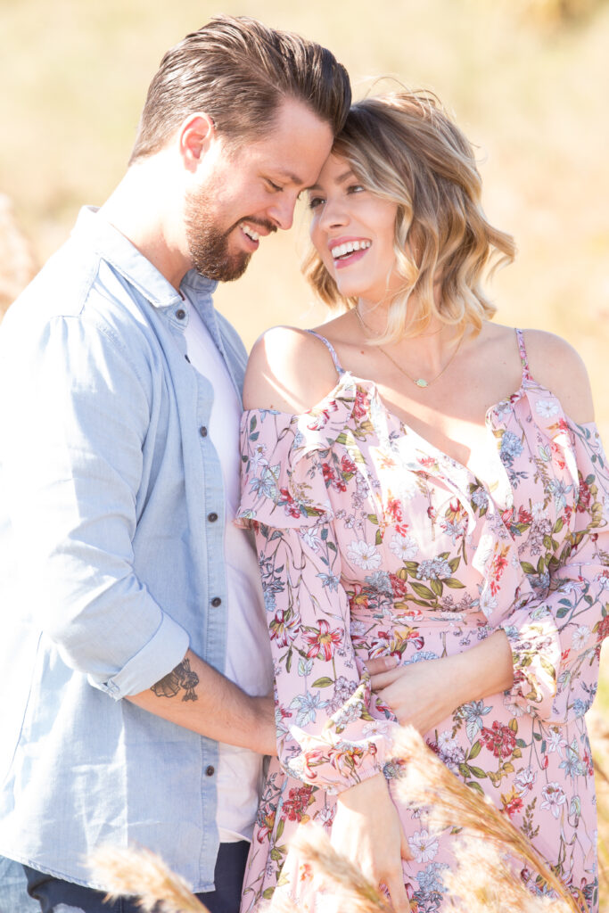 engagement photoshoot with a couple wearing light colored outfits; the man is in a light blue long-sleeve denmin shirt with a white T-shirt underneath. The woman is wearing a pink off-the-shoulder floral dress
