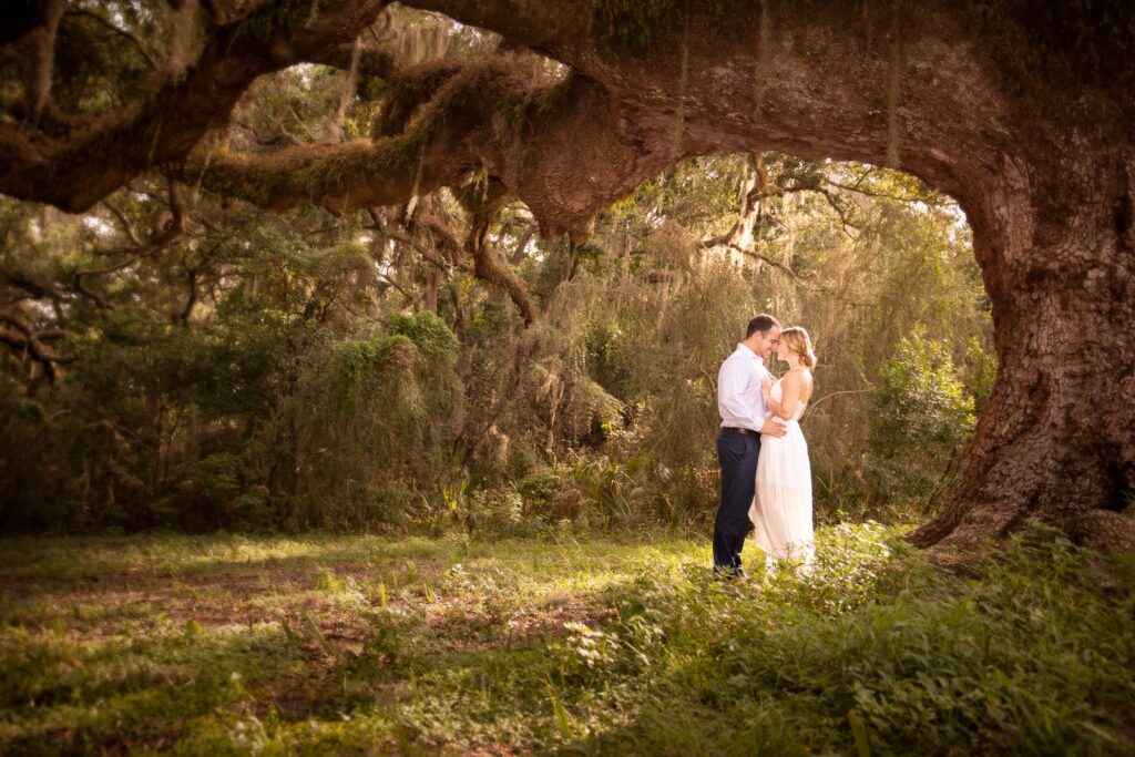 man and woman standing underneath a willow tree for the engagement photoshoot; both are wearing white outfits