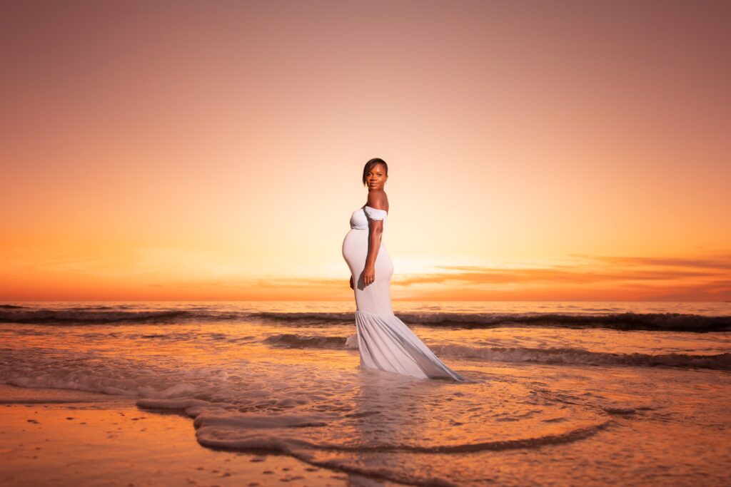 maternity photoshoot outfit ideas: black woman wearing a white bodycon mermaid dress on the shores on a Florida beach with an orange sunset in the background