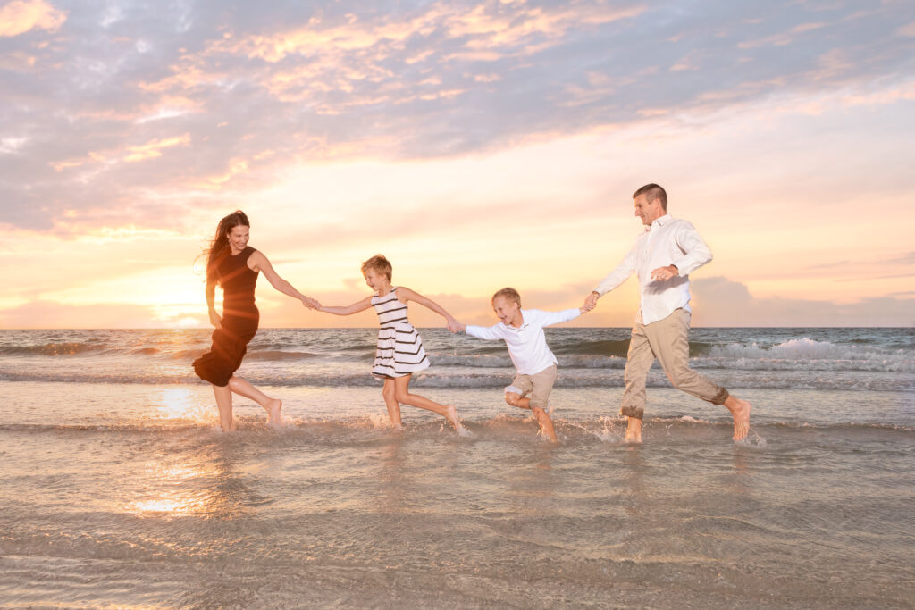 family of 4 during their beach photography session: family is running on the shores of a Tampa beach. The sun is pink, blue, and orange